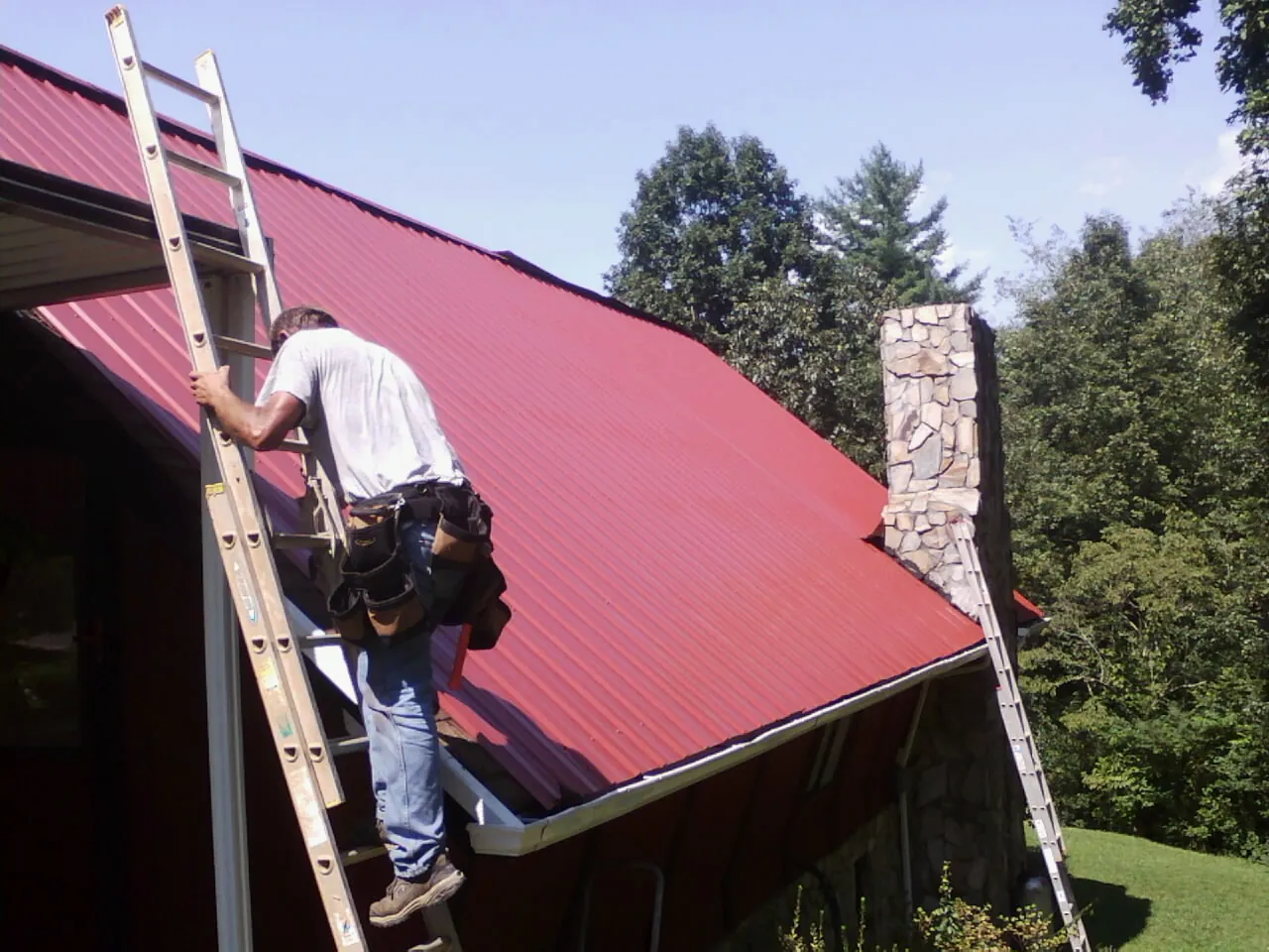 Roofer on a ladder beside a red metal roof and stone chimney — Soffit & Fascia Repair by Roofing Brothers in Grand Rapids